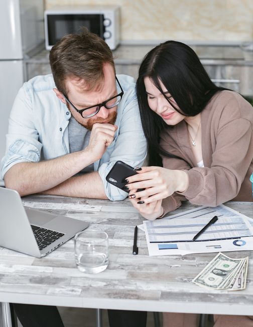 A couple examines financial papers and a smartphone at a kitchen table with a laptop.